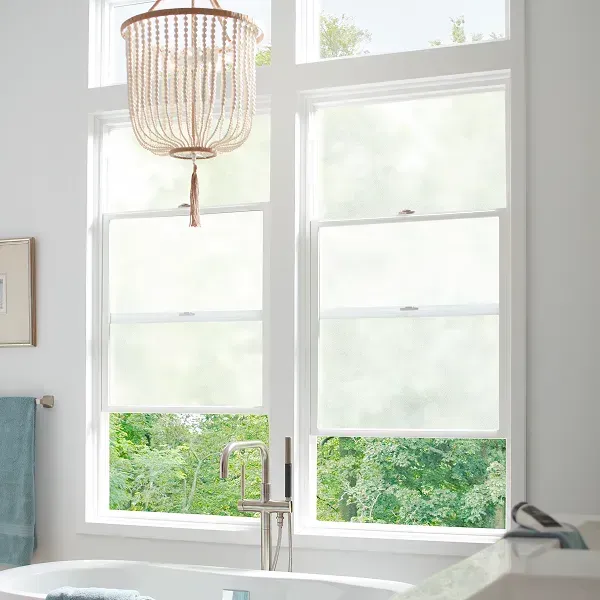 Bright bathroom with white windows, woven pendant light, and a freestanding tub by greenery outside.