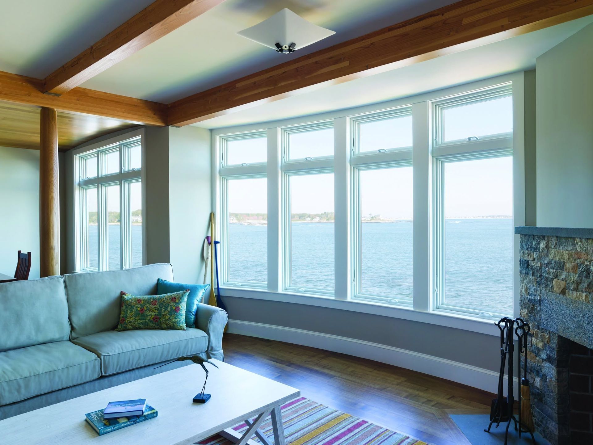 Bright living room with large bay windows, wood beams, and a light blue sofa overlooking the water