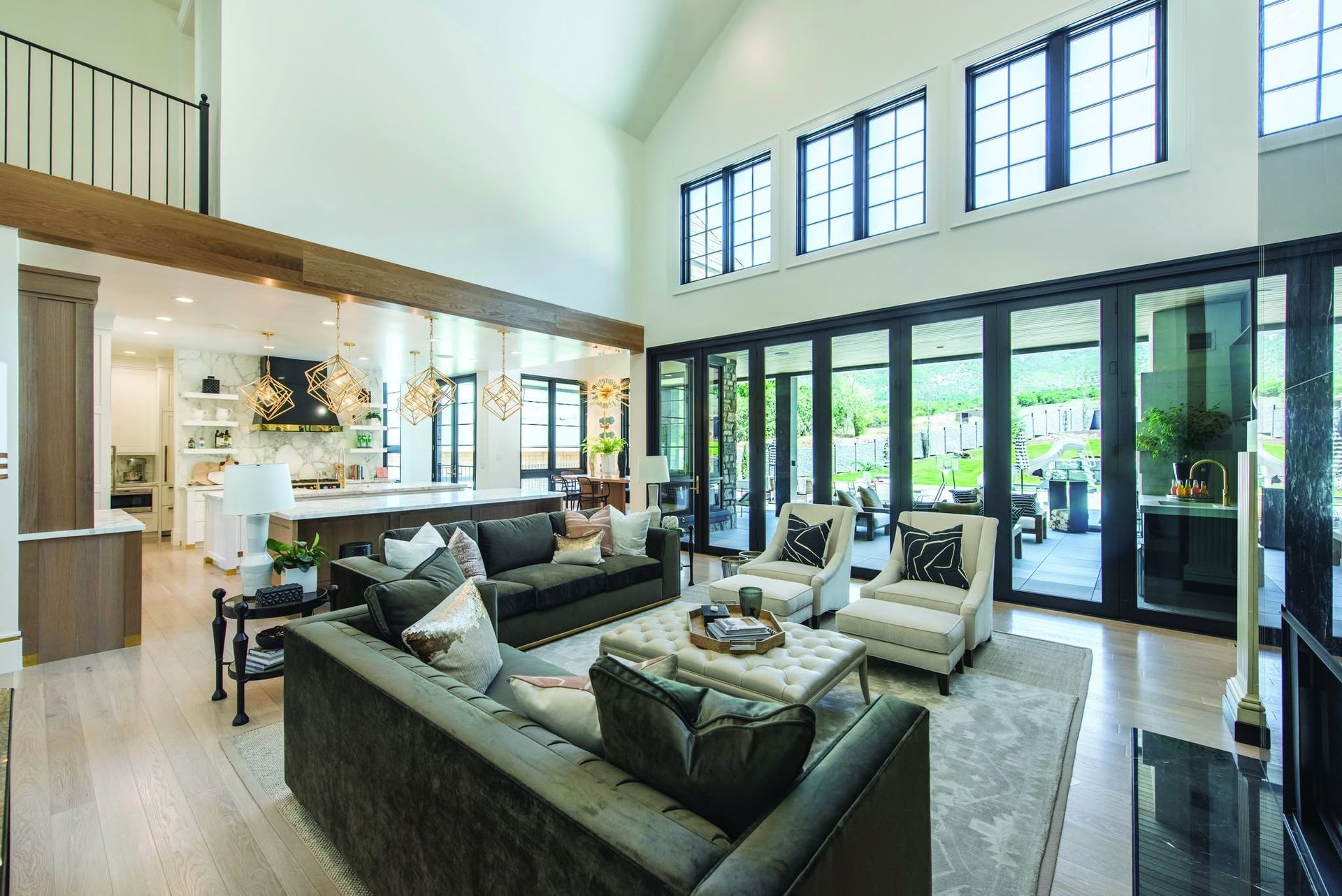 Modern open-plan living room with gray sectional, black chairs, and floor-to-ceiling glass doors overlooking greenery