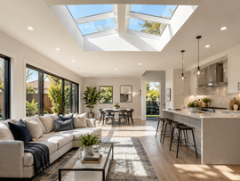 Bright modern open-plan living room and kitchen with skylights, gray sofa, dining table, and island stools.