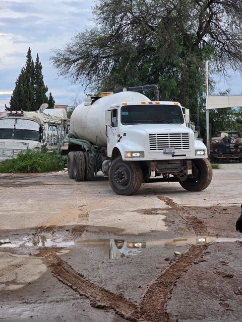 Camión cisterna de agua blanca sobre una superficie fangosa, estacionado cerca de edificios y árboles bajo un cielo nublado.