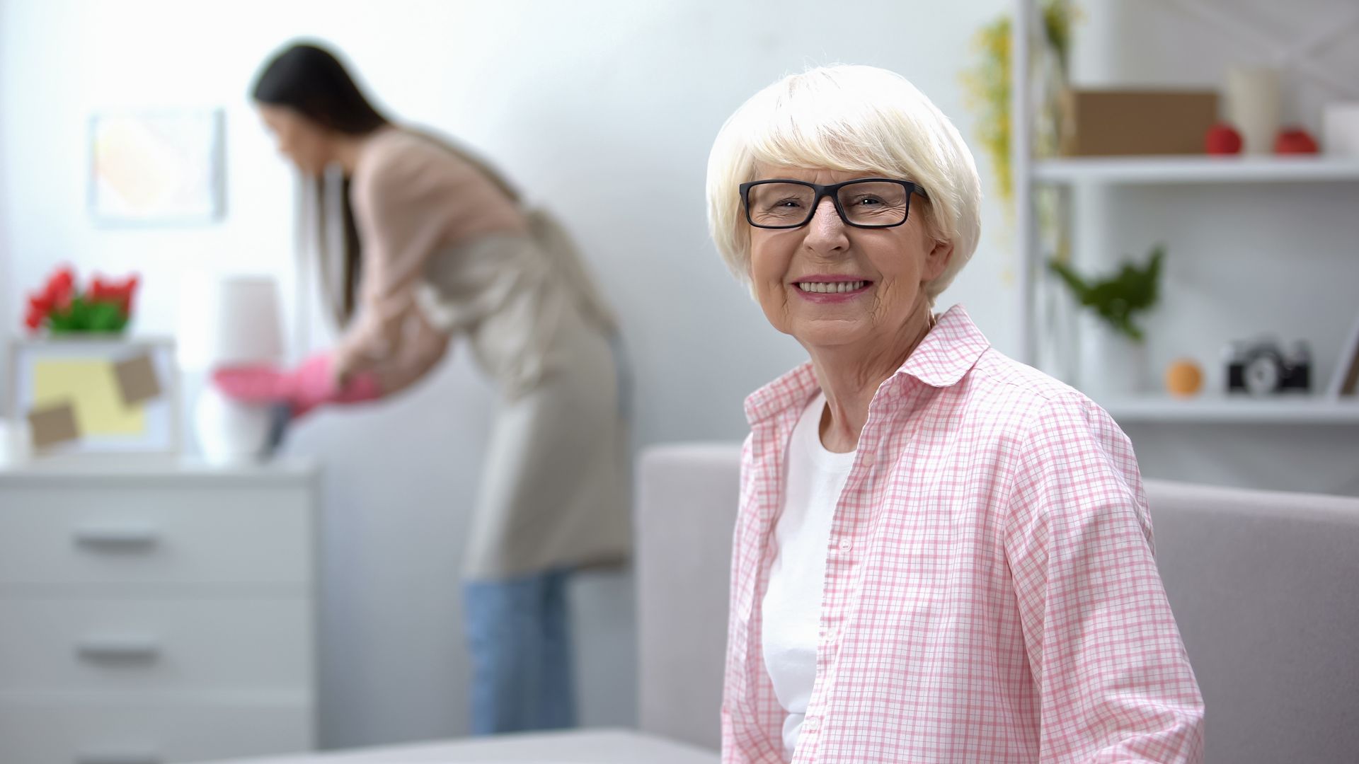 Donna sorridente con maglietta rosa e occhiali, con un addetto alle pulizie sullo sfondo che spolvera uno scaffale.