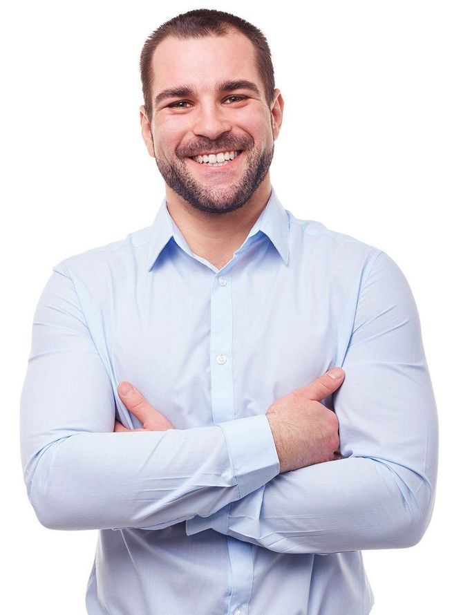 A man in a light blue shirt is standing with his arms crossed and smiling.