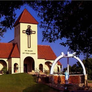 Church with red-tiled roof and cross tower beside a white arch fountain in a landscaped courtyard