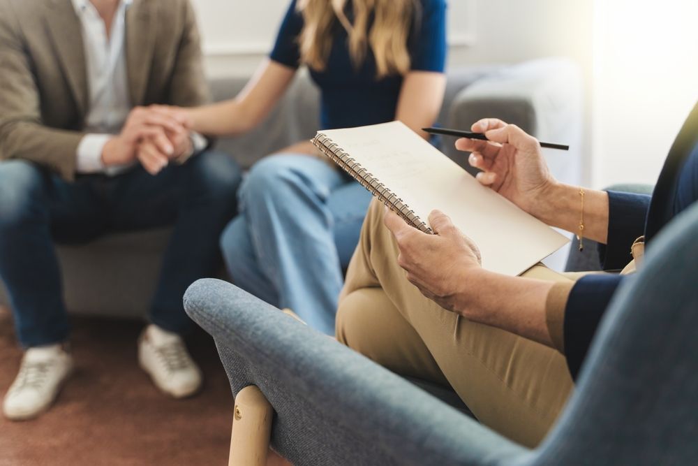 Therapist taking notes during a counseling session with a couple holding hands on a couch.