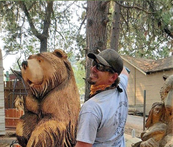 Man smiling beside a large carved bear statue outdoors in a wooded yard