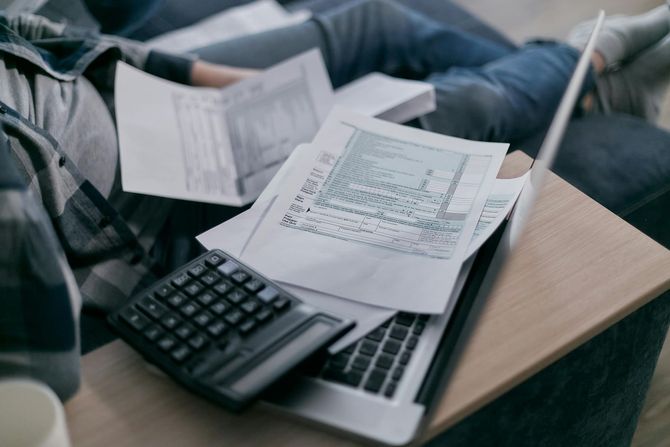 Person working on taxes with calculator, papers, and laptop on lap.