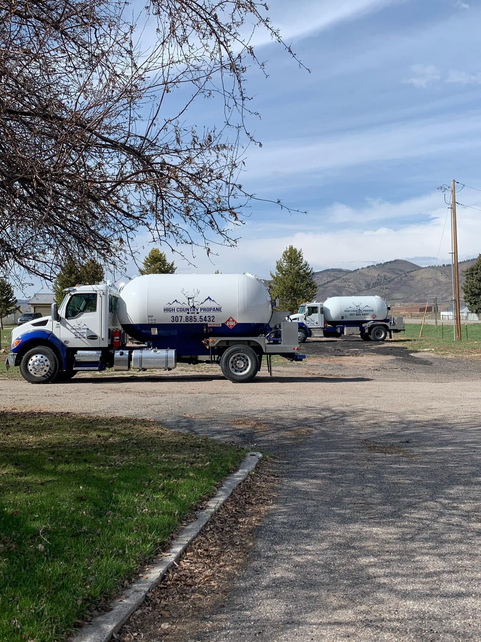 Two white utility trucks on a gravel lot near trees and mountains under a cloudy sky.