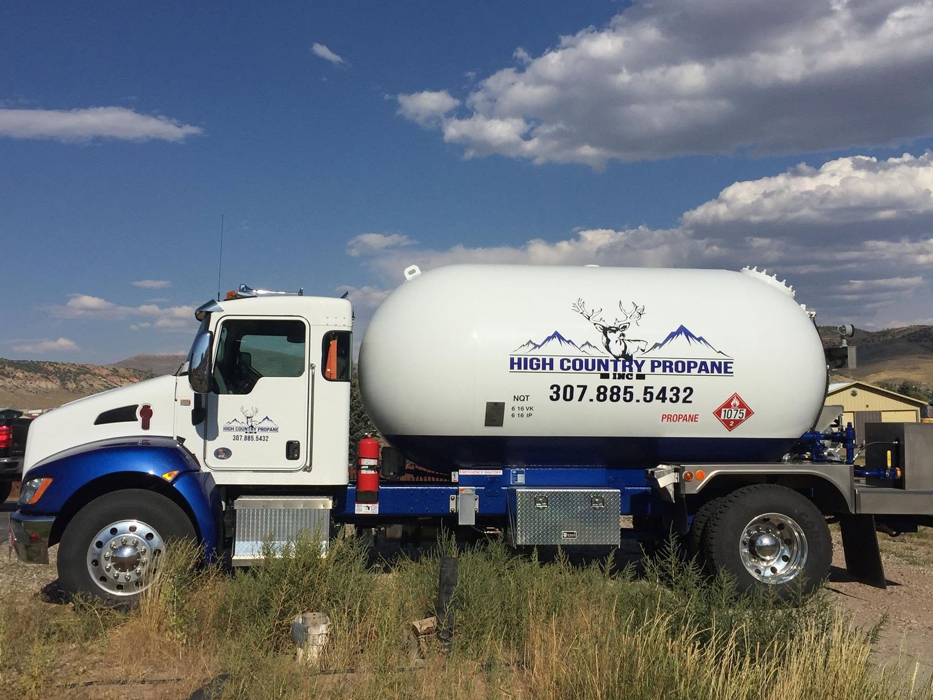 White and blue water tanker truck on a grassy roadside under a cloudy blue sky