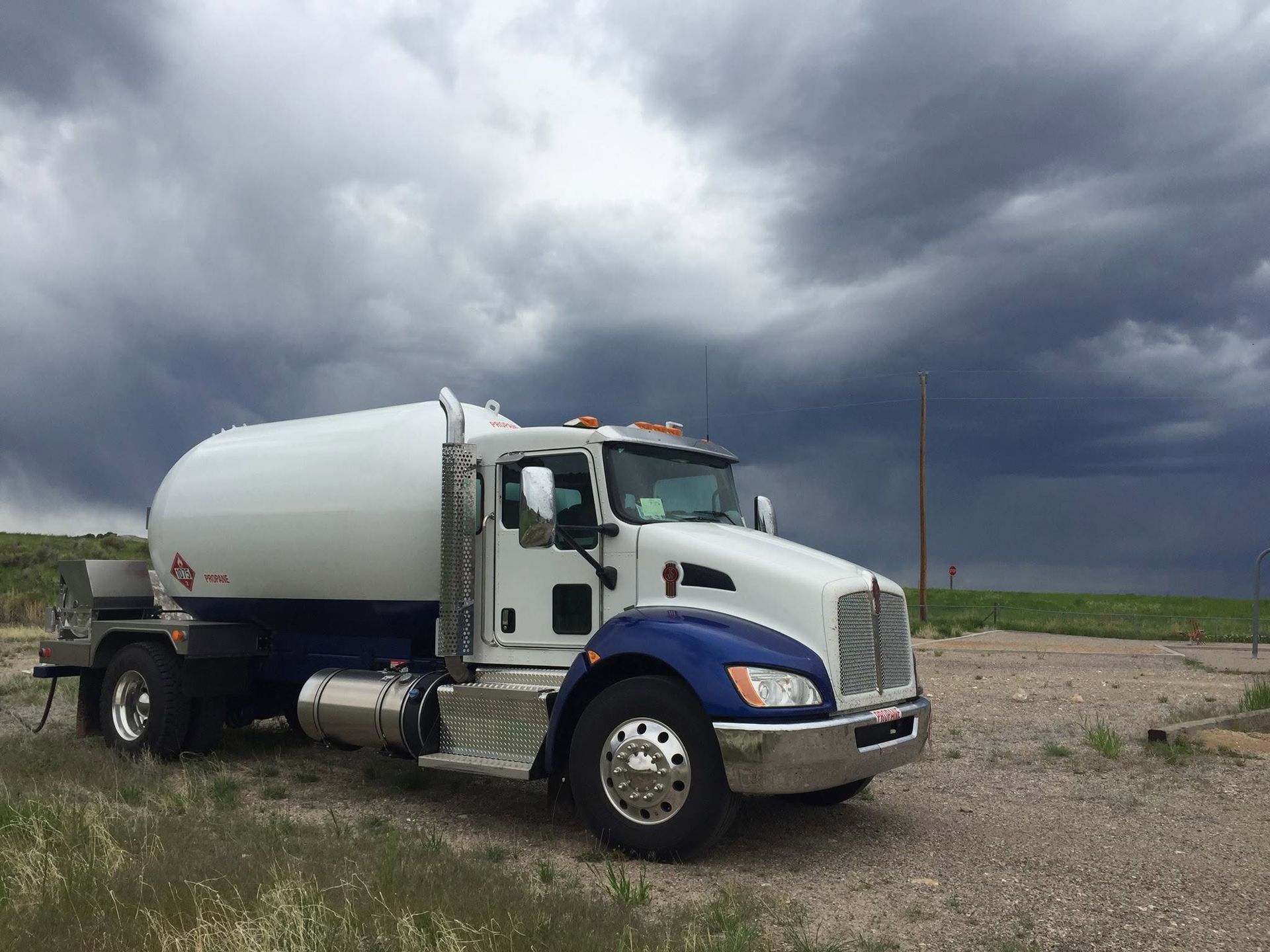 White and blue tanker truck parked on gravel under dark storm clouds