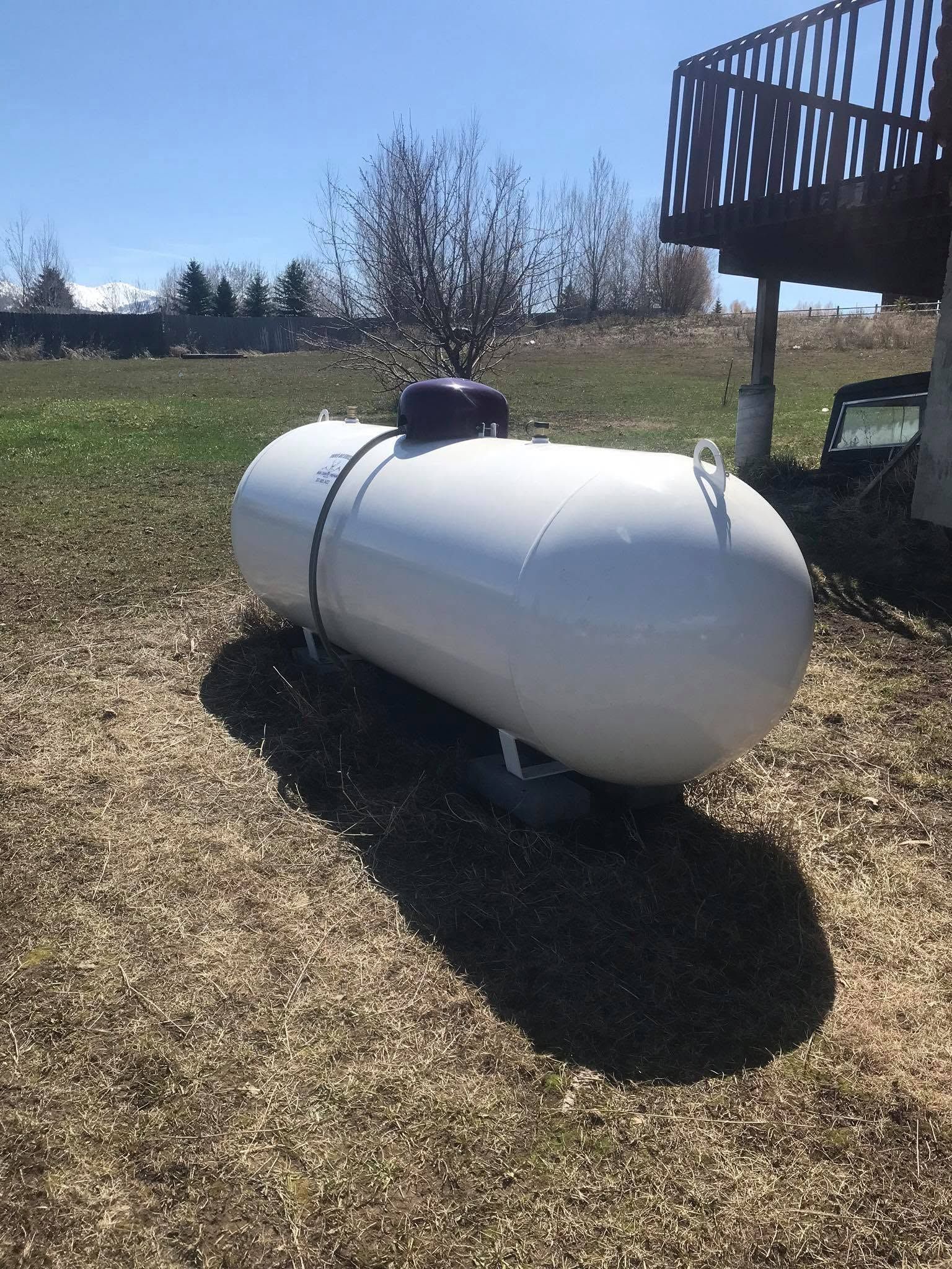 White propane tank on gravel beside a wooden deck in a grassy yard under a clear sky