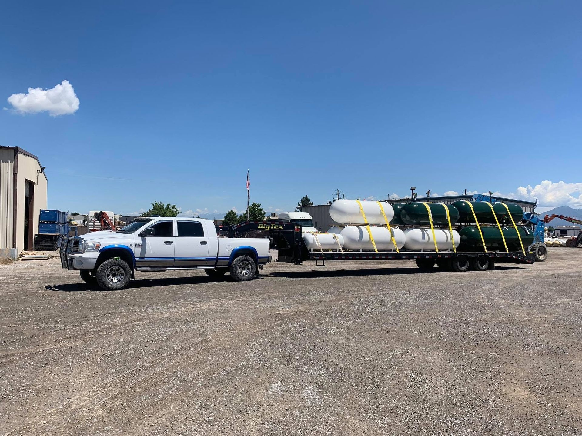 White pickup truck hauling large cylindrical tanks on a trailer in a gravel lot under a blue sky