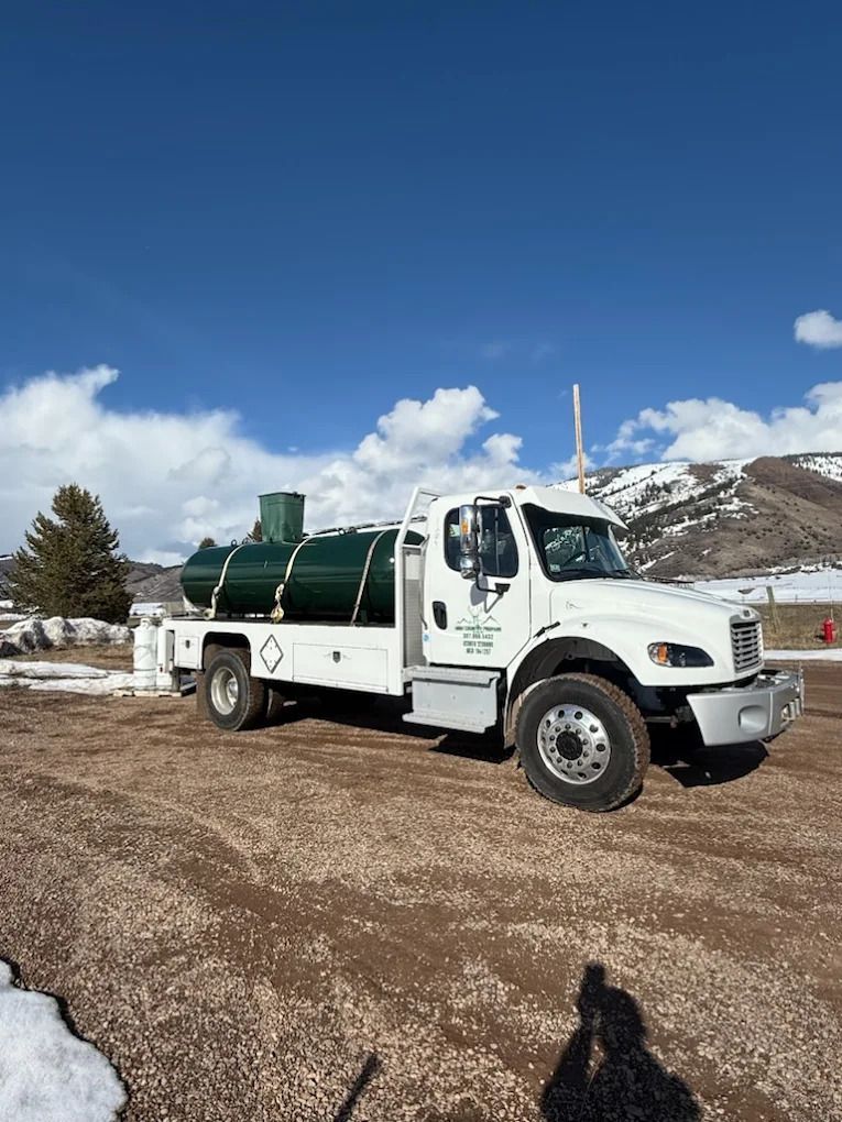 White utility truck with green tank parked on a gravel lot beside snowy mountains under a blue sky