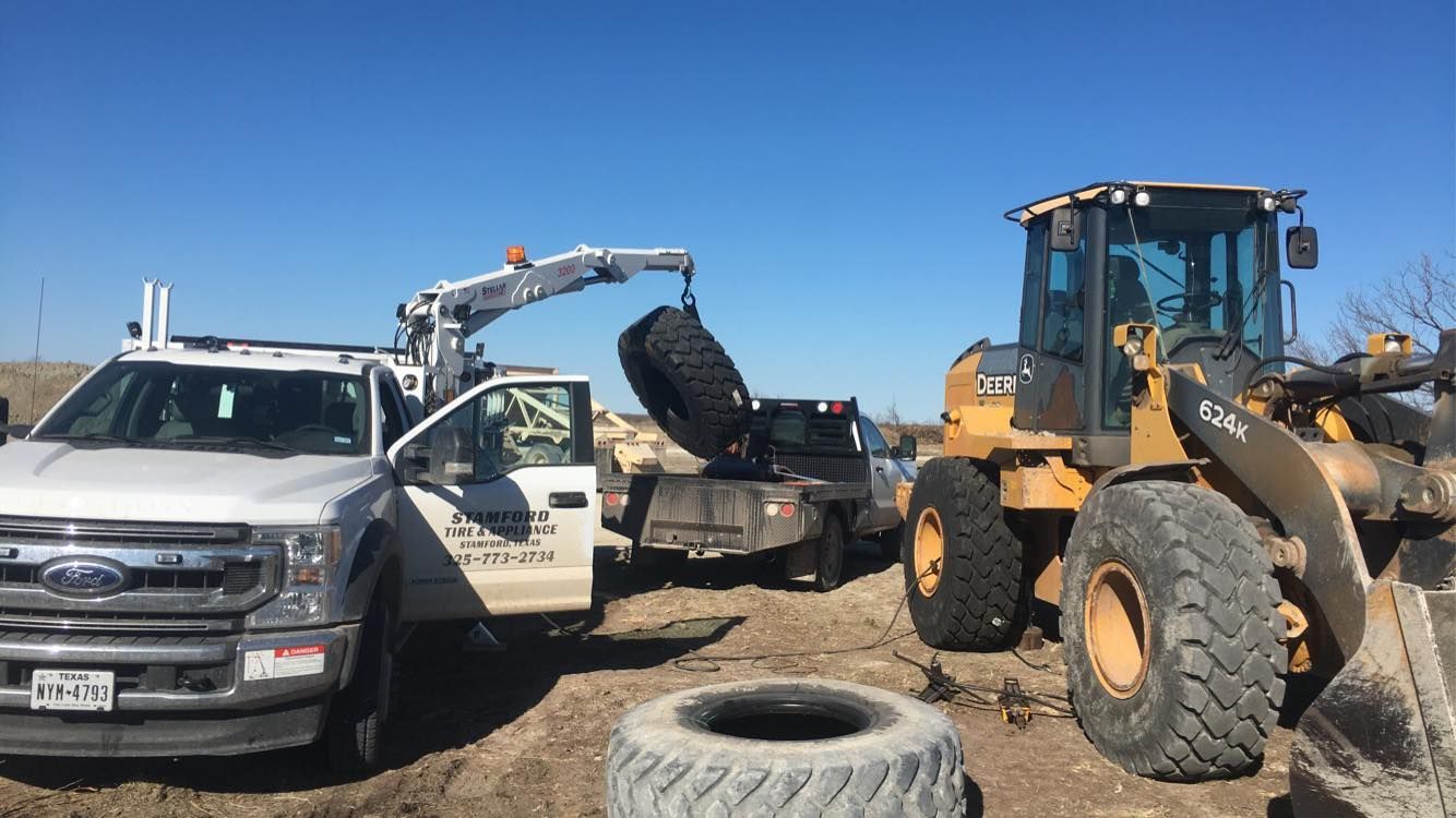 Tow truck and front loader beside a white pickup in a dusty lot, with a tire in the foreground.
