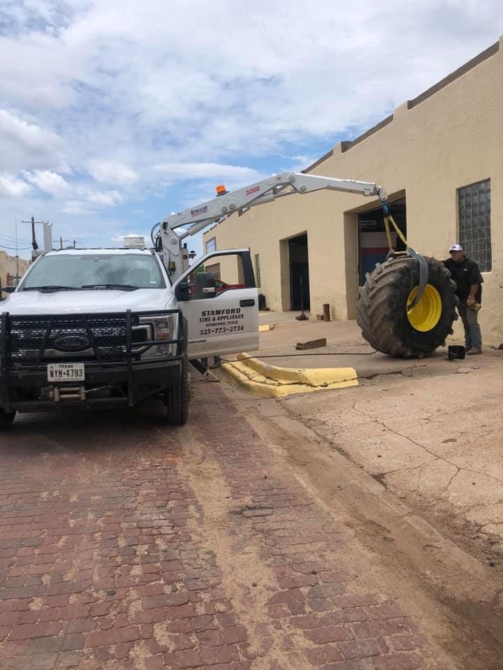 White truck with boom lift beside a beige building and a large tractor on a brick street