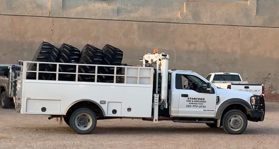 White utility truck loaded with black tires, parked on a dirt road beside a wall.