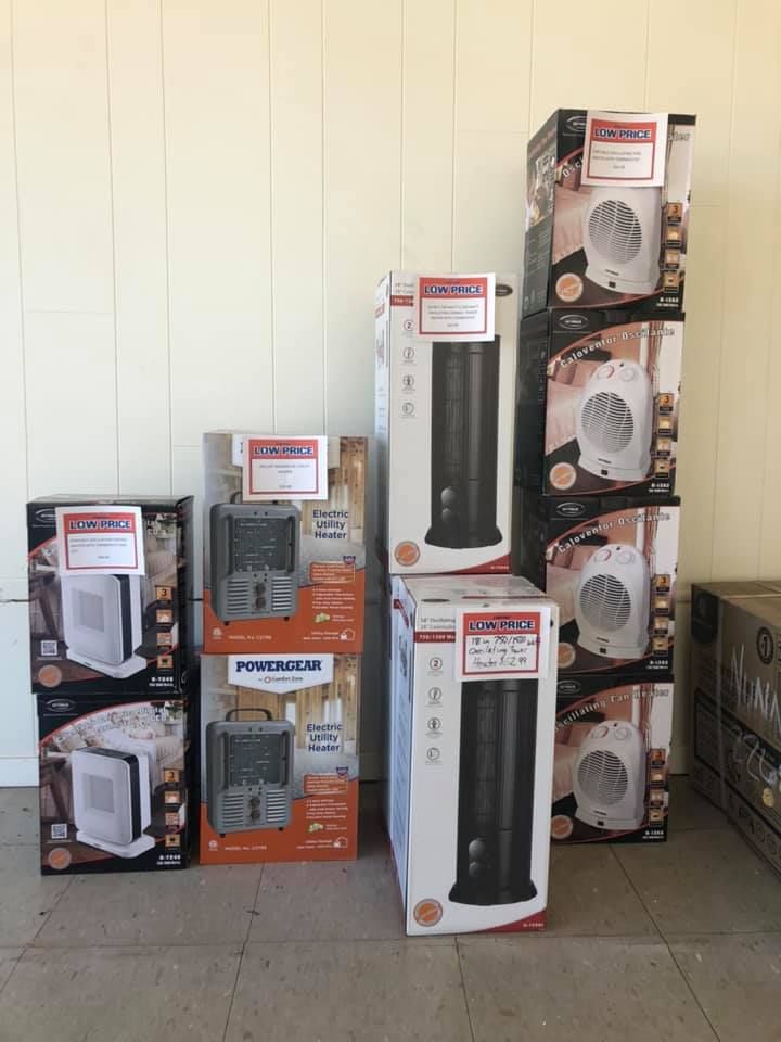 Stacked boxed household fans and space heaters displayed indoors against a white wall.