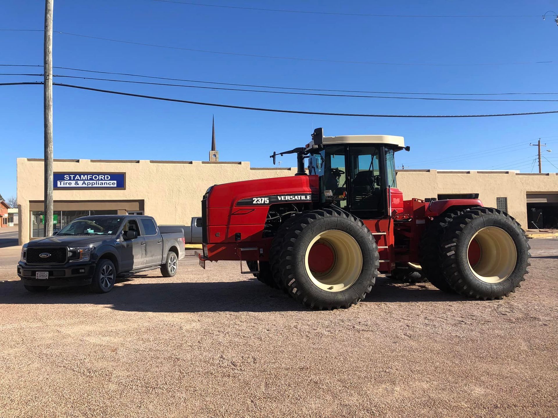 Red tractor parked beside a gray car in front of a beige building under a clear blue sky