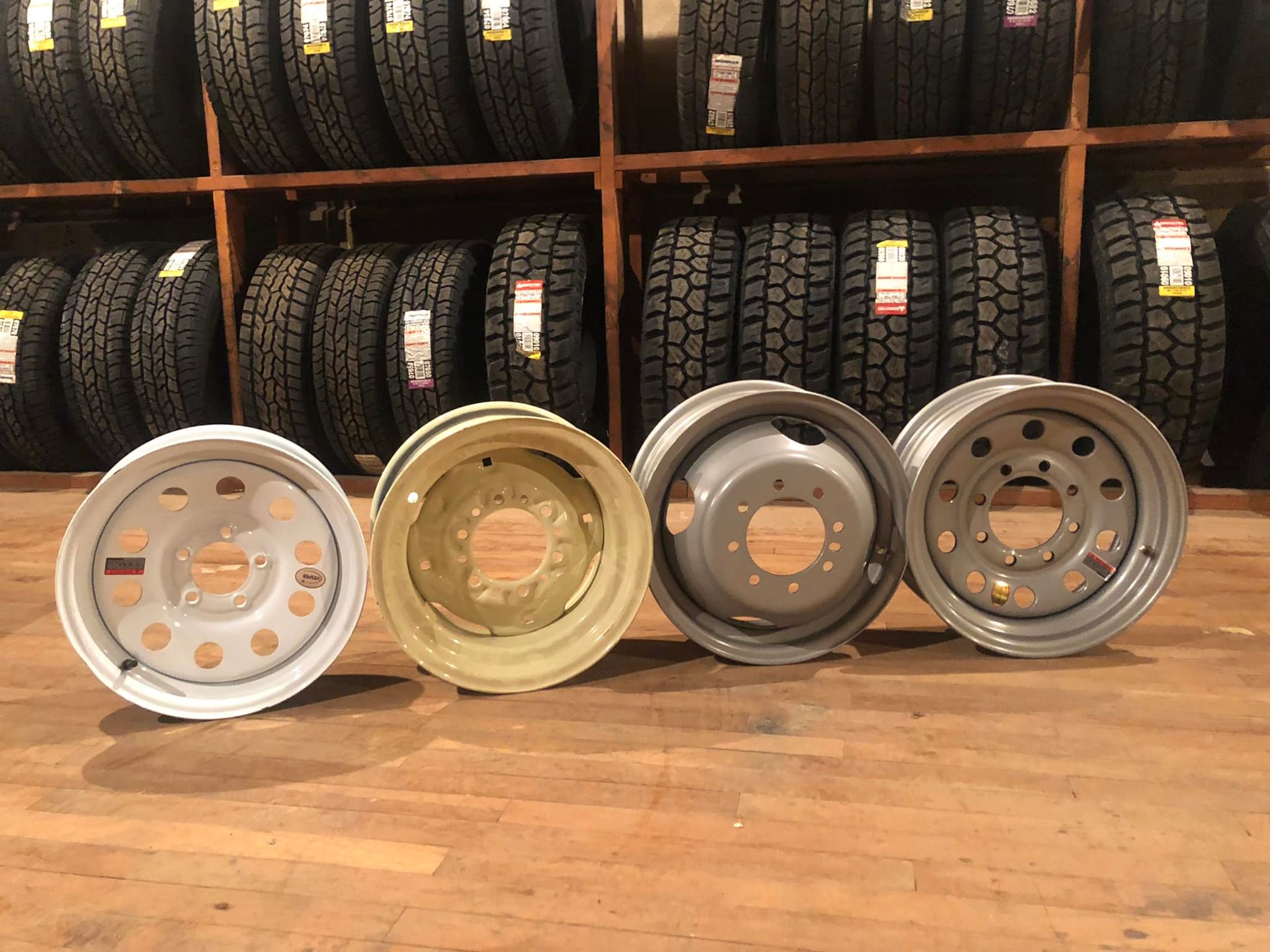 Four alloy rims on a wooden floor in front of stacked tires on shelves.