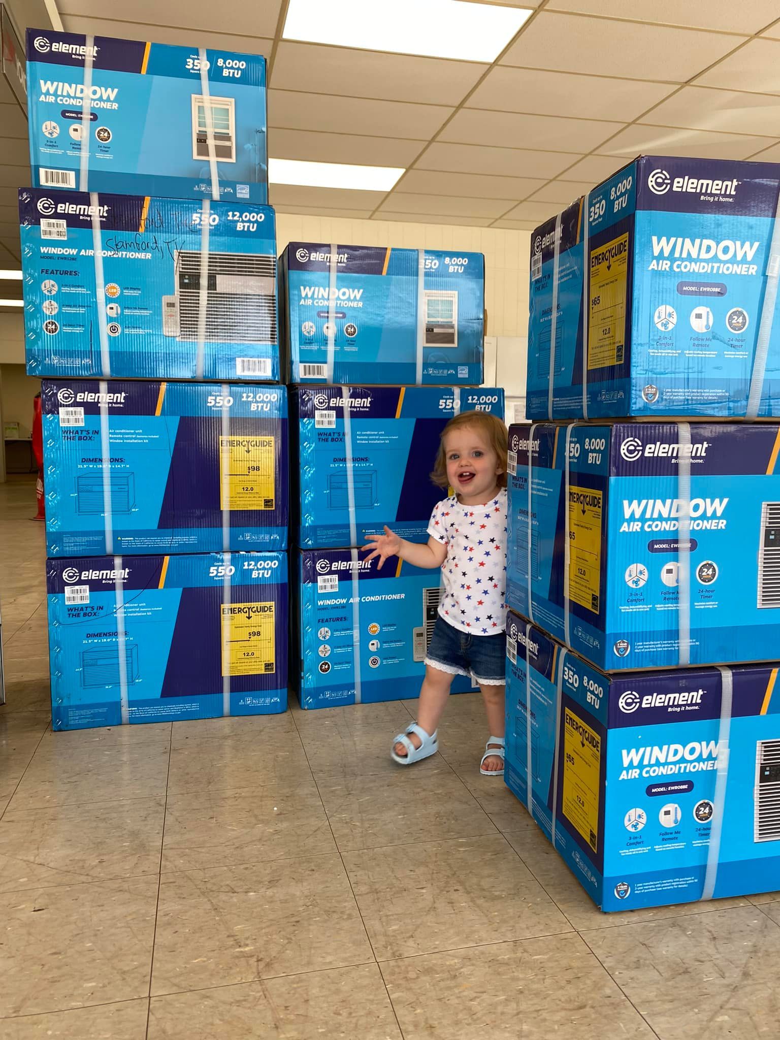 Young child standing between stacks of blue boxed fans in a store aisle, smiling and pointing.
