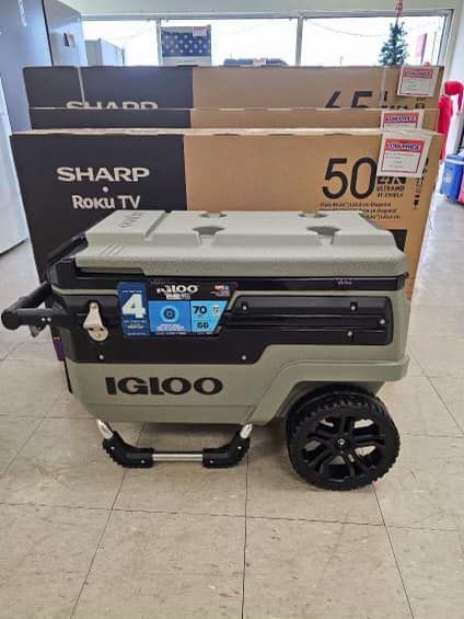 Igloo cooler on wheels with a grill on top, in front of stacked TV boxes in a store.