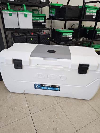 White Igloo cooler on a tiled floor in front of stacked black storage bins and boxes.