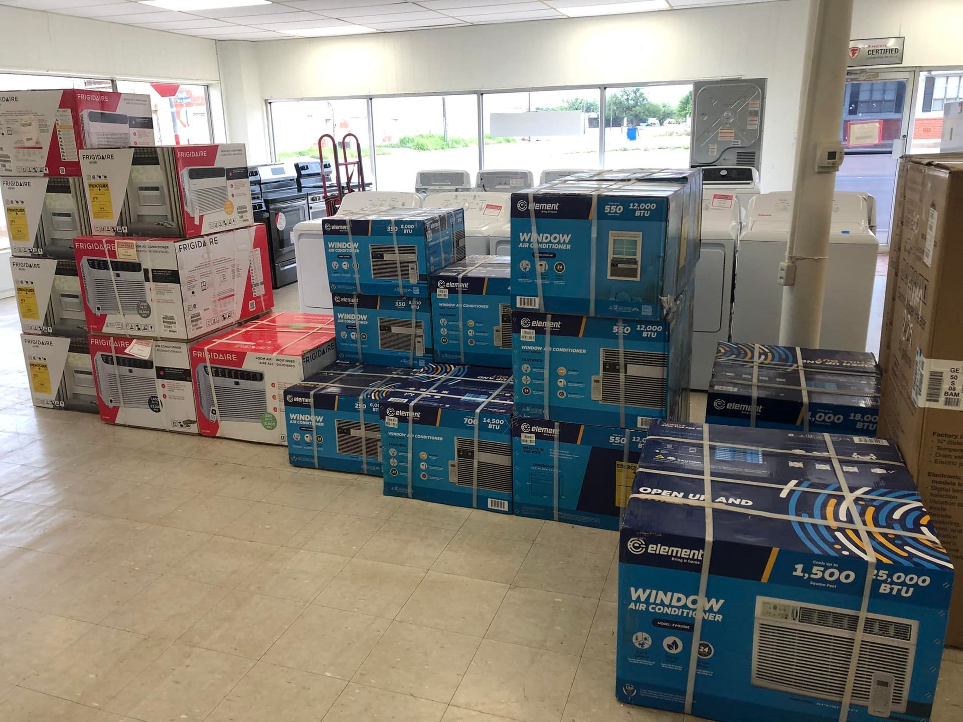 Stacks of boxed air conditioners and appliances in a store aisle near a bright front window.