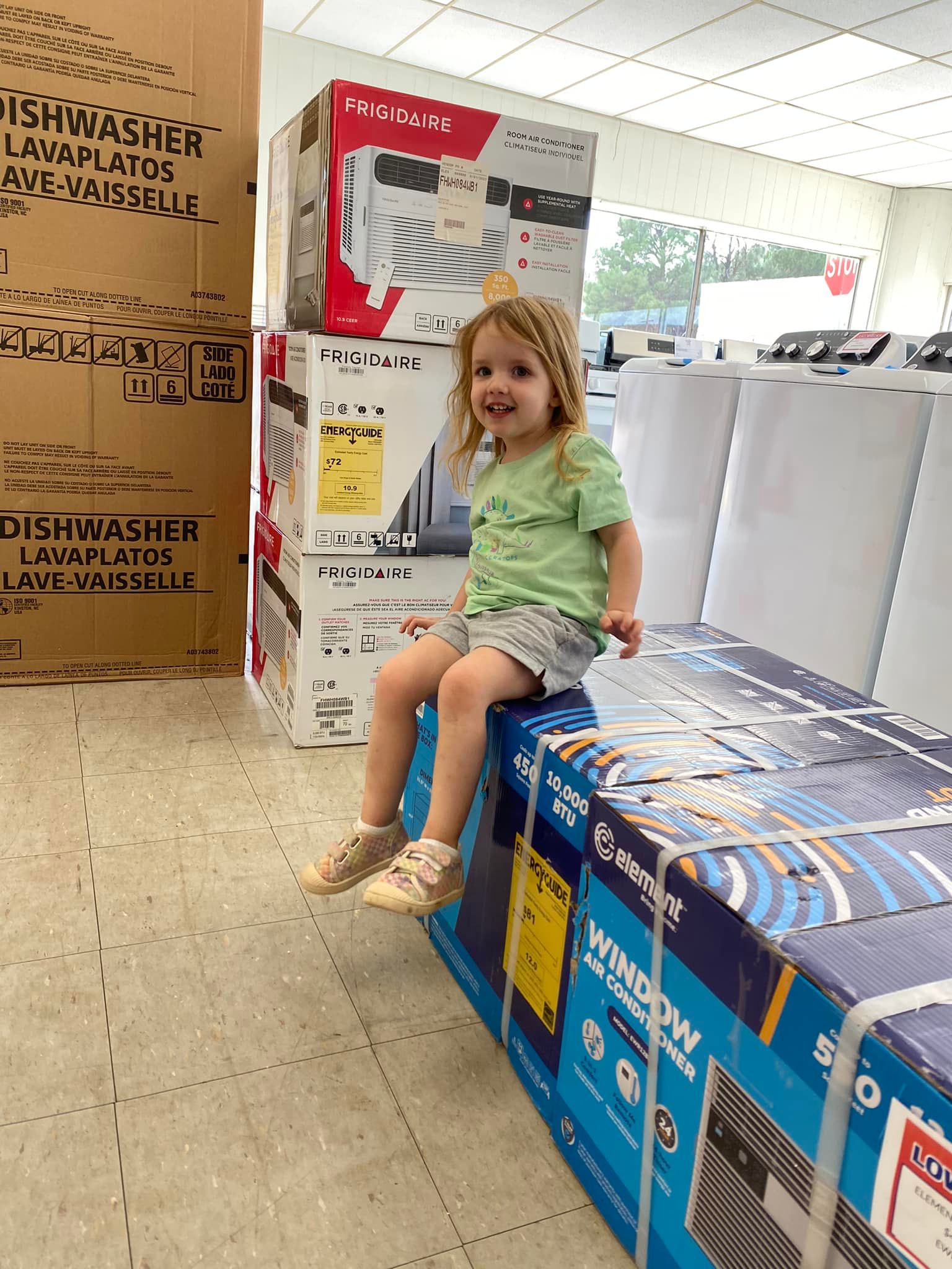 Child sitting on stacked appliance boxes in a store aisle, smiling at the camera.