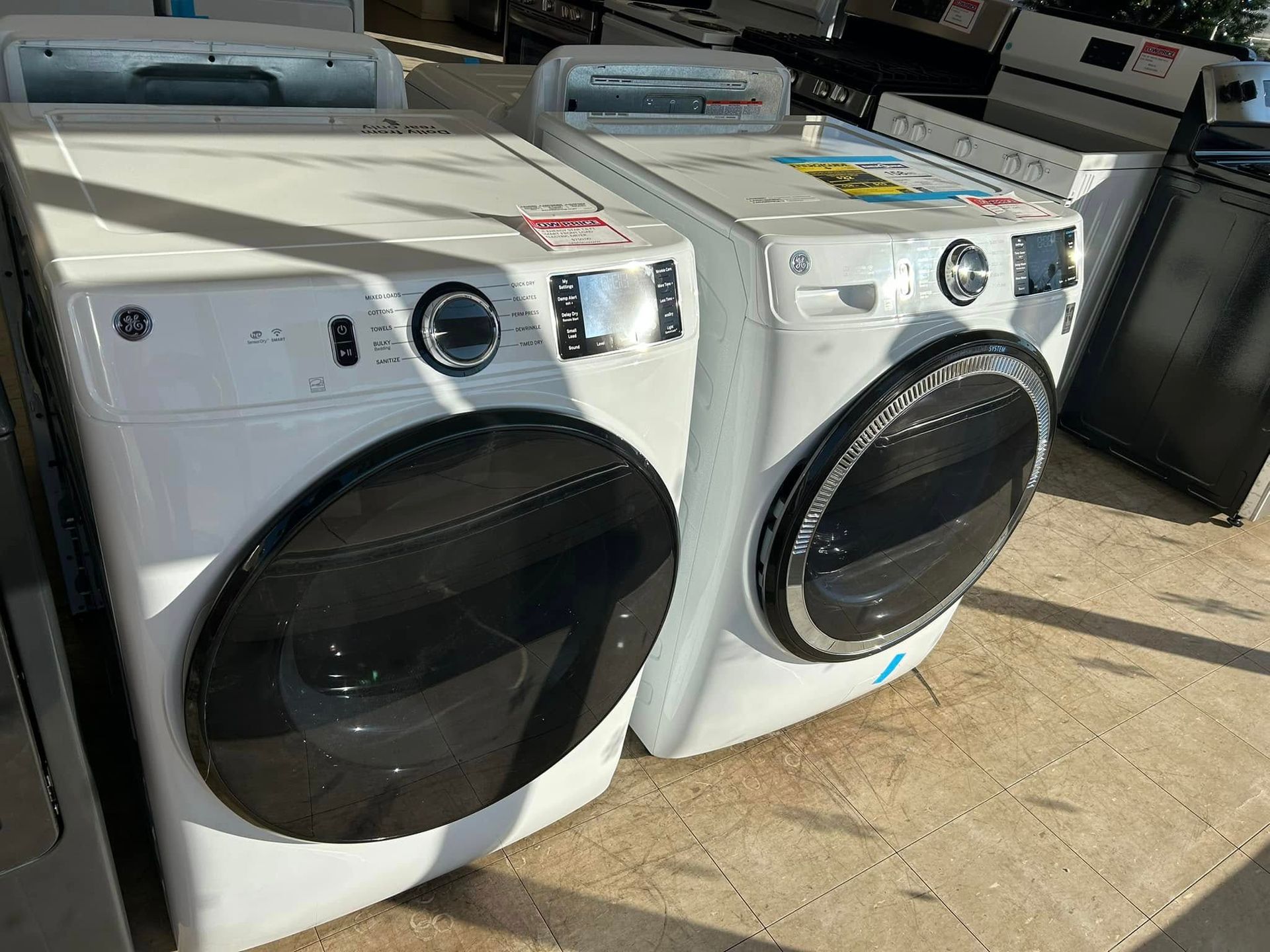 Two white front-load washers with black doors, lined up outdoors in a sunlit appliance lot.