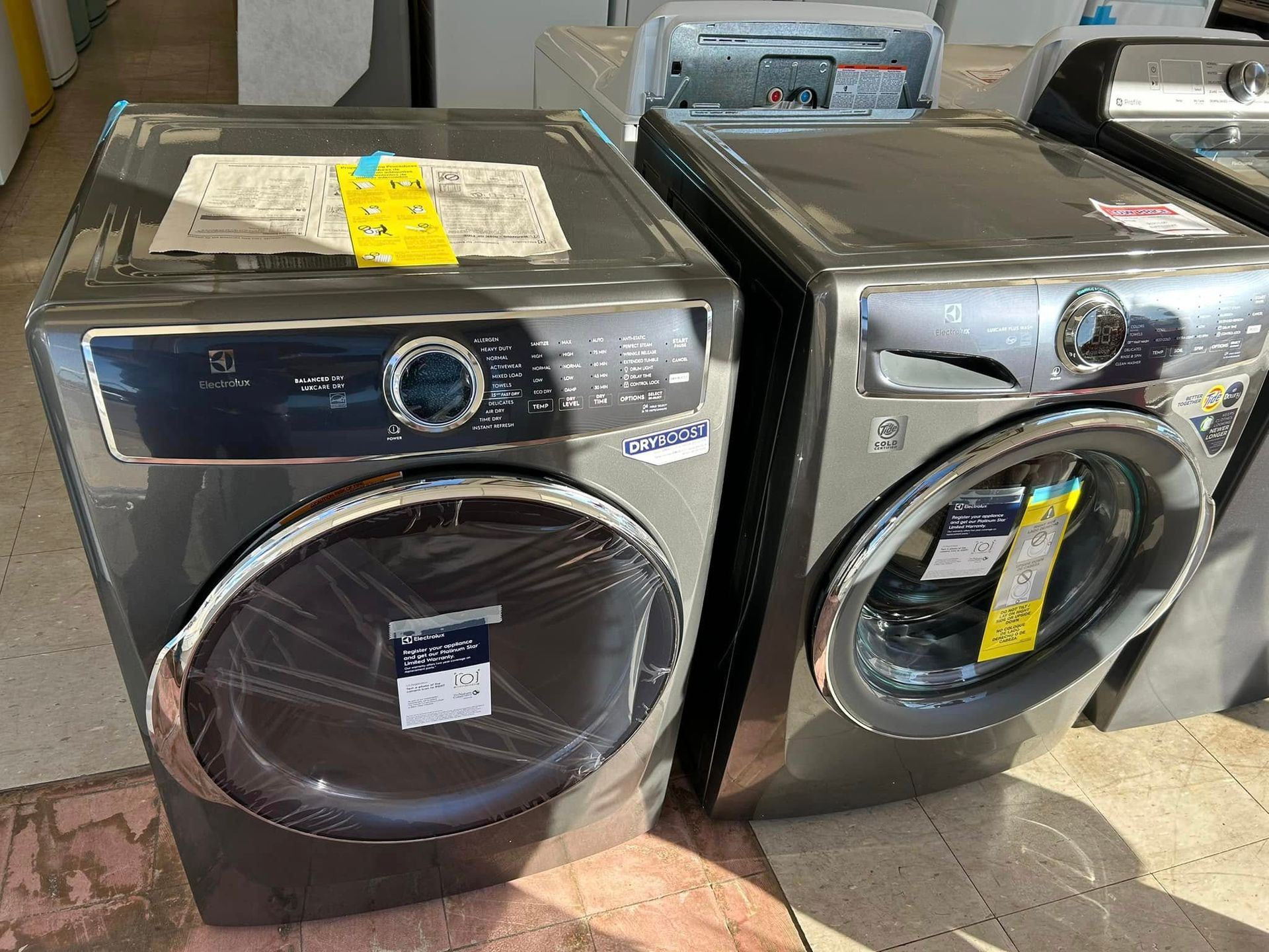 Two front-load washers in a store, one black and one silver, wrapped and tagged.