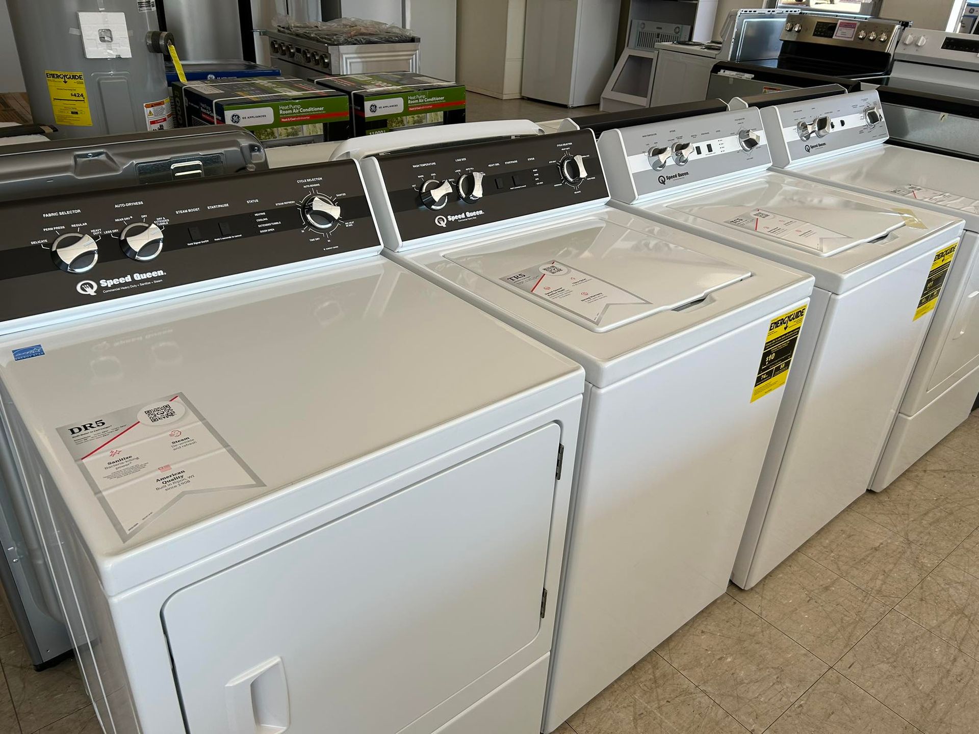 Row of white and black laundry machines in a laundromat or appliance showroom.