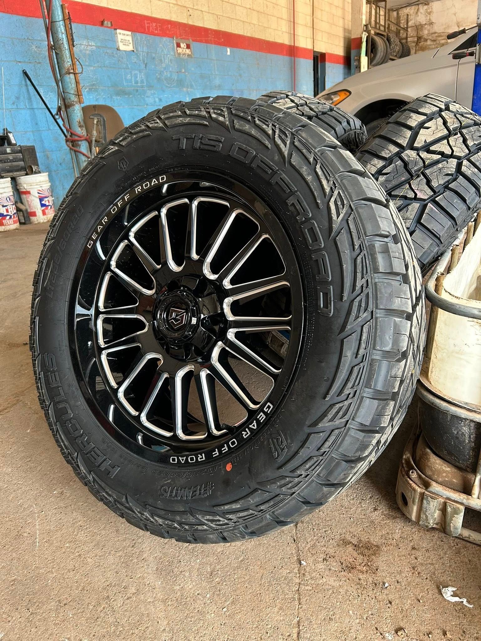 Black off-road tire with glossy black rim leaning in a garage, with another tire behind it.