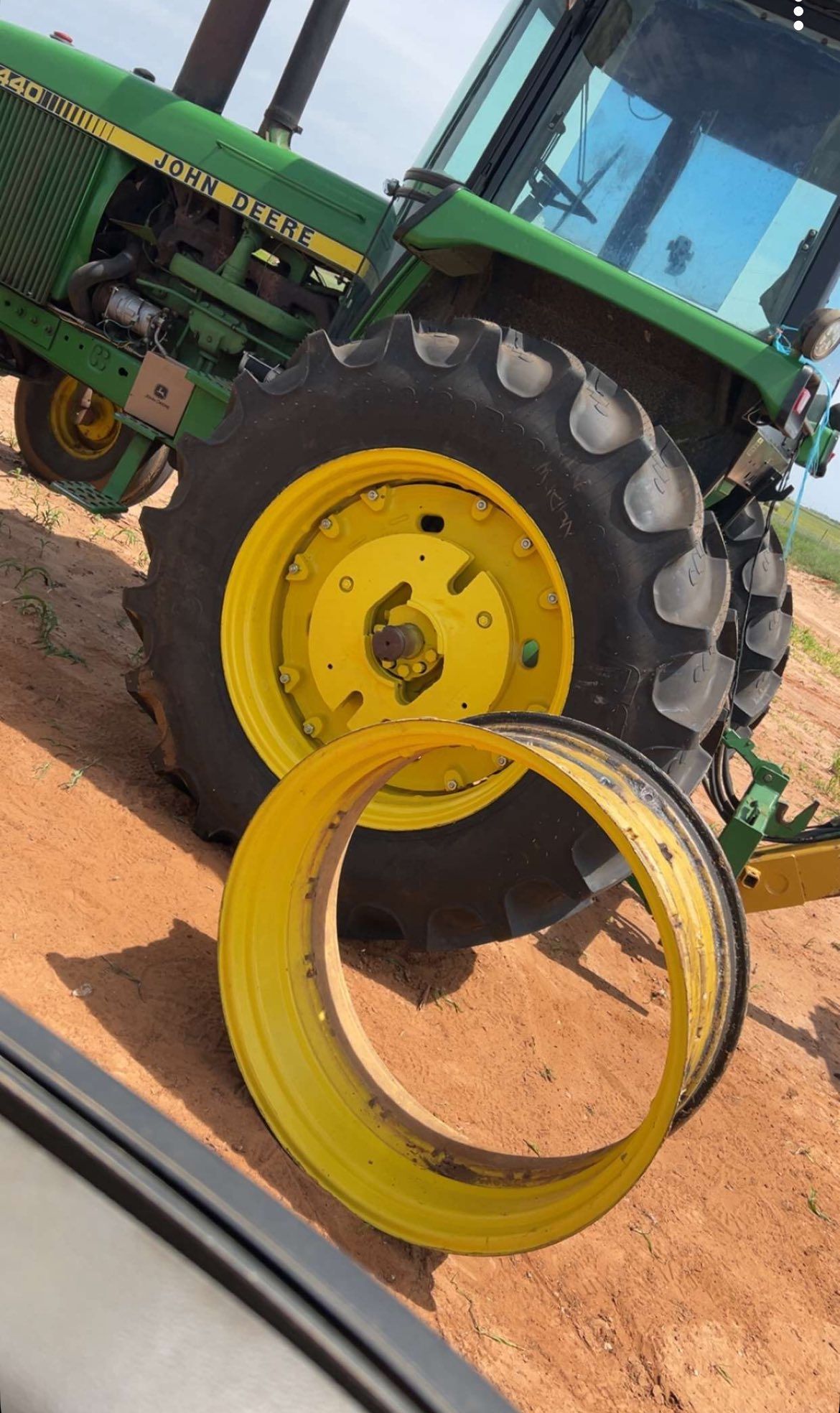 Green tractor with yellow wheel and detached tire rim on sandy ground, outdoors in sunlight