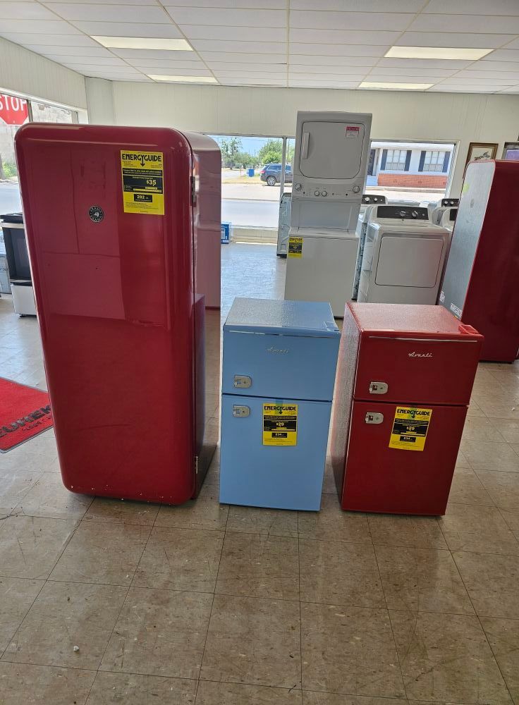 Three retro refrigerators in red, blue, and red in a store aisle with a gray appliance behind them