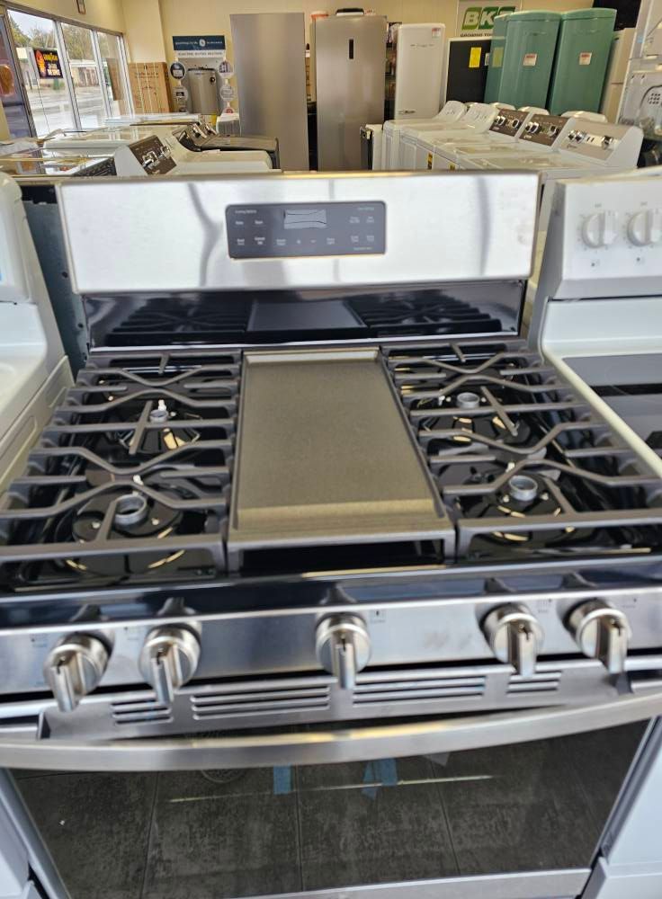 Stainless steel gas range with six burners and center griddle, displayed in an appliance store.