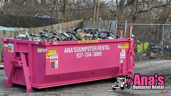 Bright pink Ana’s Dumpster Rental dumpster filled with debris beside a chain-link fence outdoors