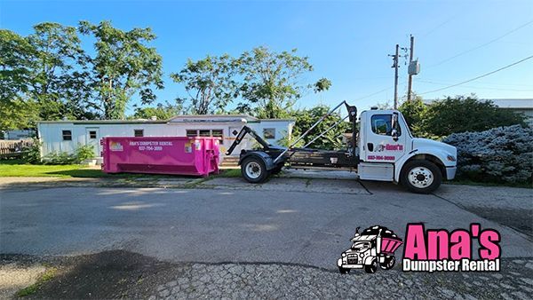 White tow truck hauling a pink dumpster on a sunny street, with Ana's Dumpster Rental logo in the corner
