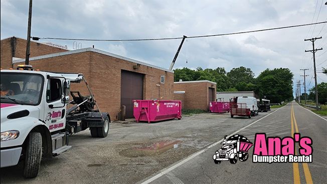 Dumpsters lined beside a brick building, with a white truck parked on a roadside under cloudy skies.