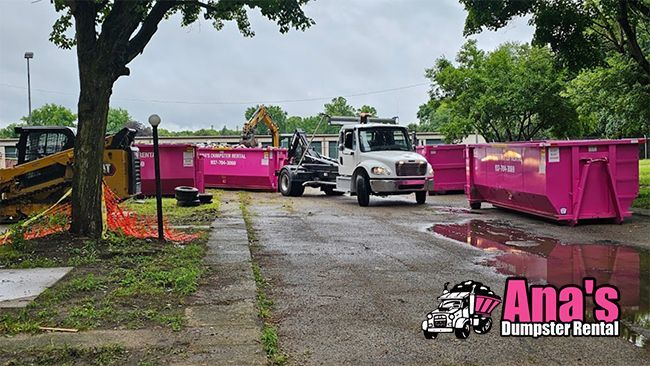 Magenta dumpsters lined up beside a driveway with a white truck and a worker in the background