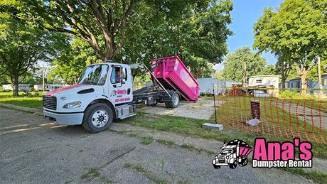Pink dump truck tipping on a street beside a white tow truck in a leafy neighborhood.