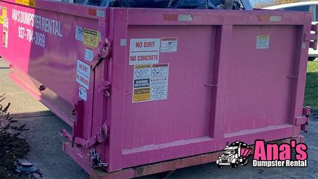 Pink dumpster with warning stickers in an outdoor parking area, marked