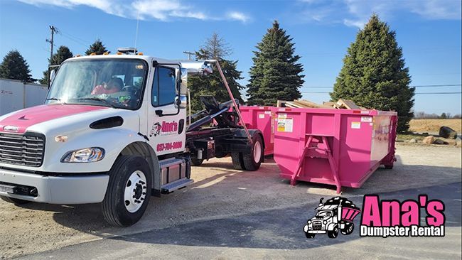 White tow truck hauling a pink dumpster on a sunny lot with trees in the background