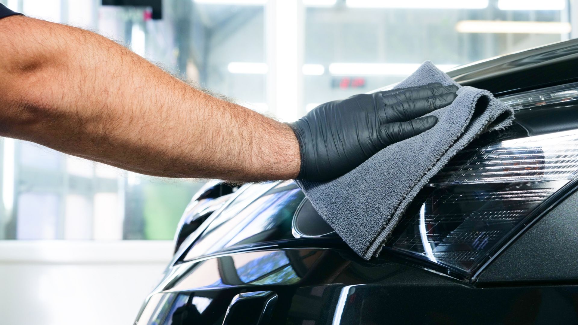 A gloved hand wiping a black car with a microfiber cloth in a garage.