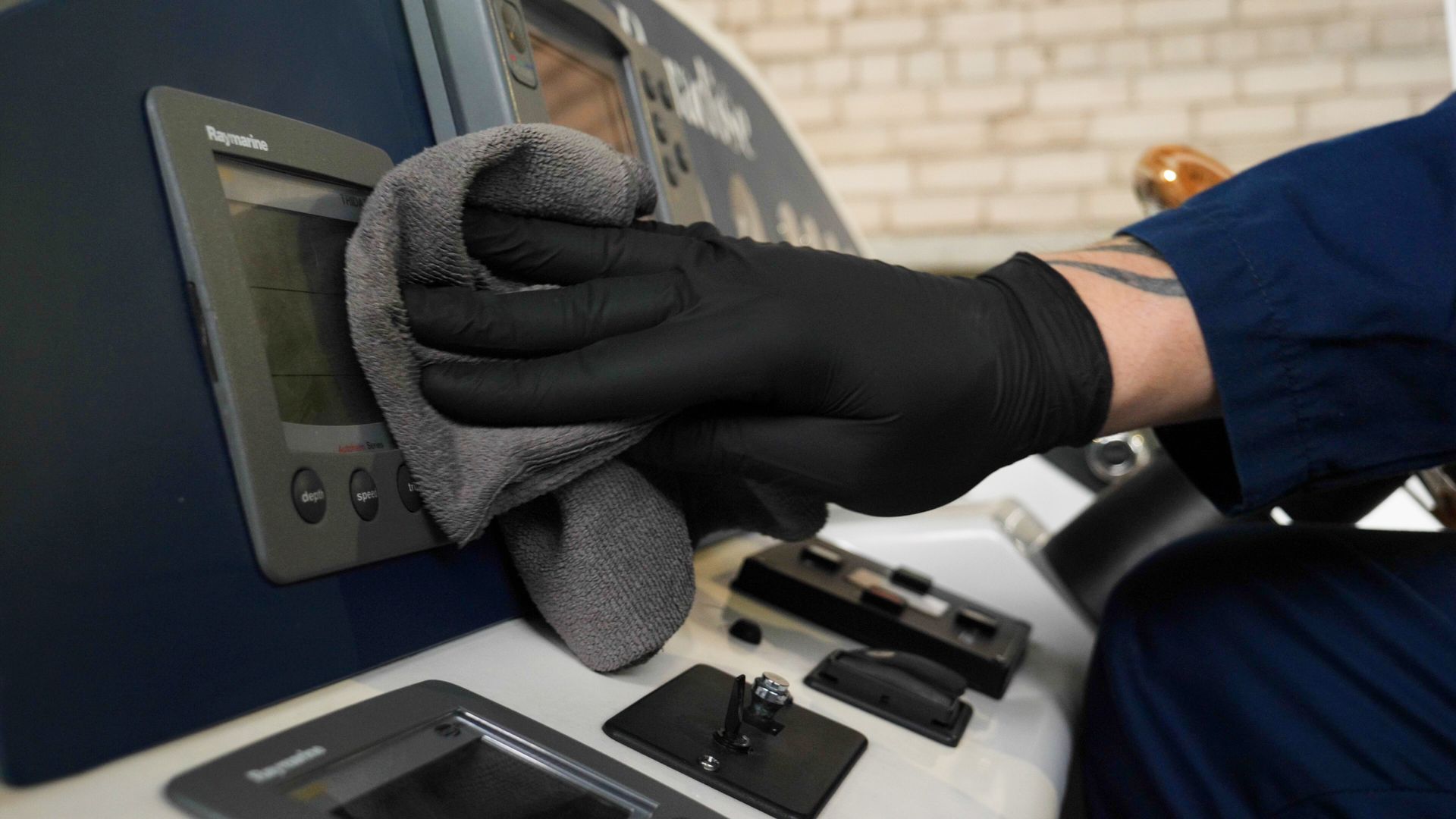 Gloved hand wiping a wall-mounted control panel with a gray cloth in a workshop setting