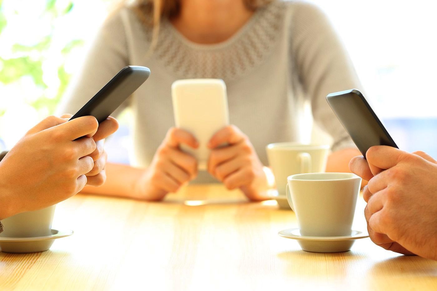 Three people at a table using smartphones with coffee cups in a bright cafe