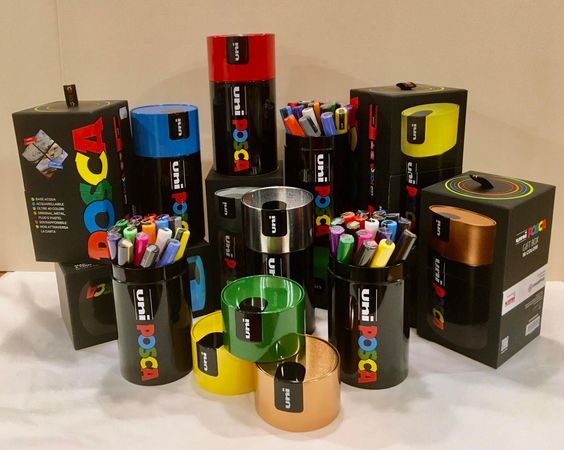 Scatole cilindriche e imballaggi assortiti e colorati con il marchio DCC, disposti su una superficie bianca.