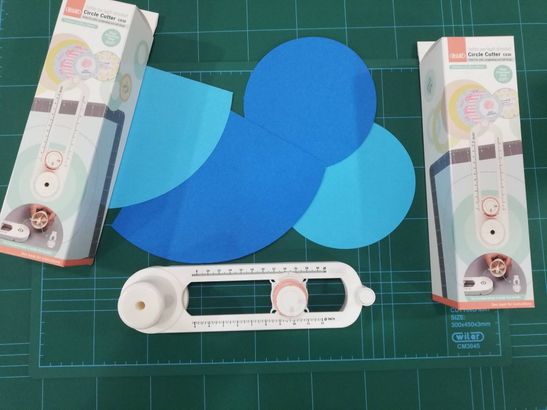 Manico bianco per taglierina artigianale con ritagli di carta circolari blu e confezione in scatola su un tappetino da taglio.