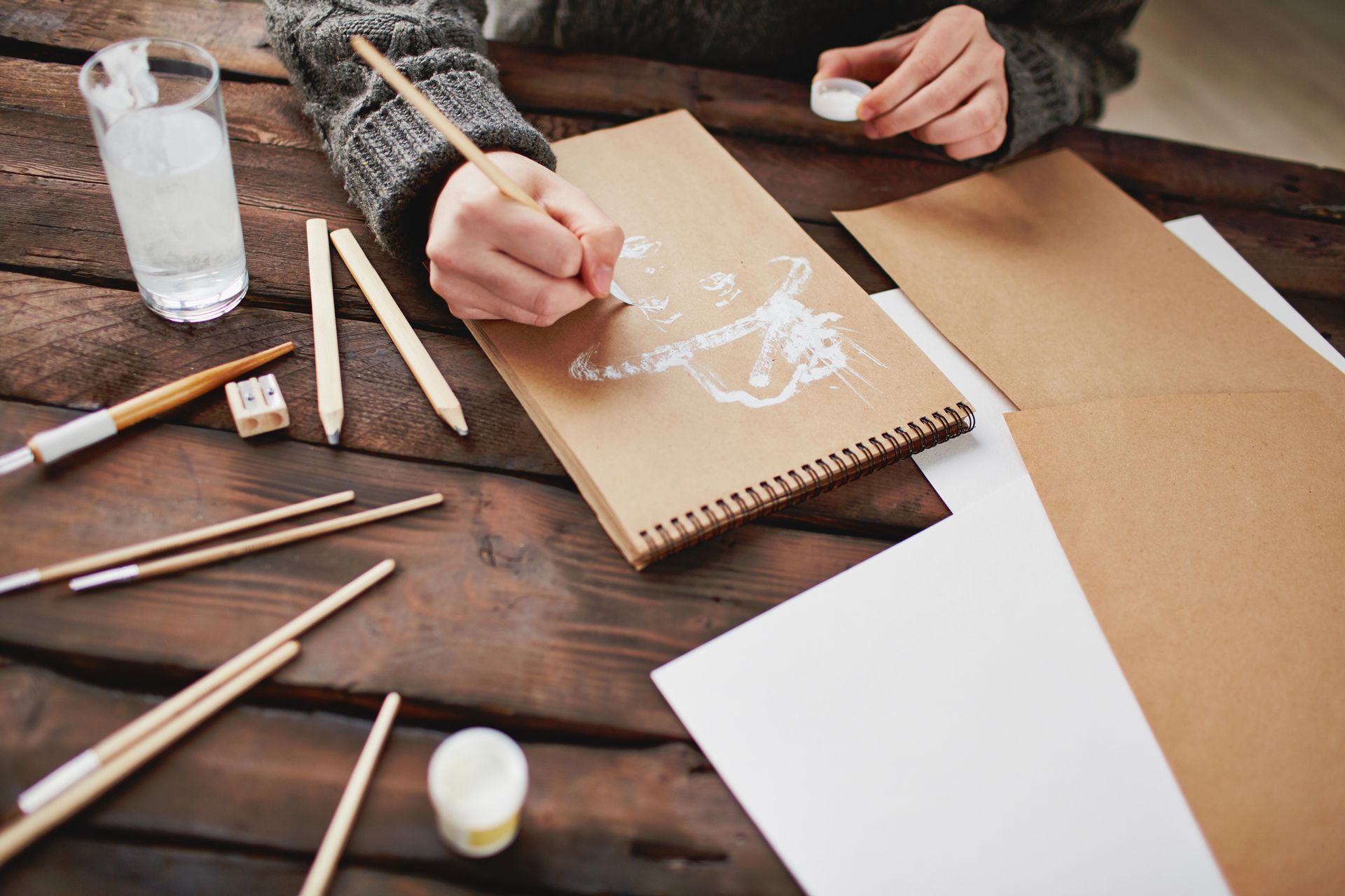 Mani che disegnano su un quaderno marrone a una scrivania di legno con matite, carta e una bottiglia d'acqua.