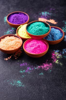 Cinque ciotole di polvere colorata su una superficie scura, con pigmenti viola, gialli, turchesi, blu e rosa sparsi tutt'intorno.