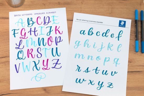 Due fogli per esercitarsi nella calligrafia dell'alfabeto con lettere corsive blu e viola su una scrivania di legno.